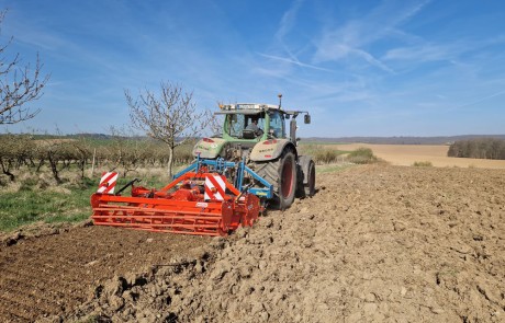 Ein Traktor pflügt ein großes, trockenes Feld unter einem blauen Himmel mit Wolkenfetzen. Bäume säumen den Rand des Feldes, und in der Ferne erstreckt sich offenes Ackerland.