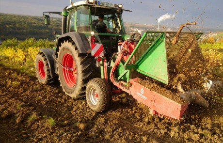 Ein grüner Traktor mit roten Rädern gräbt und hebt den Boden auf einem Feld mit Hilfe von landwirtschaftlichen Geräten, die an der Front angebracht sind. Erde und Pflanzenwurzeln fliegen in die Luft. Im Hintergrund sind üppige grüne Hügel zu sehen.