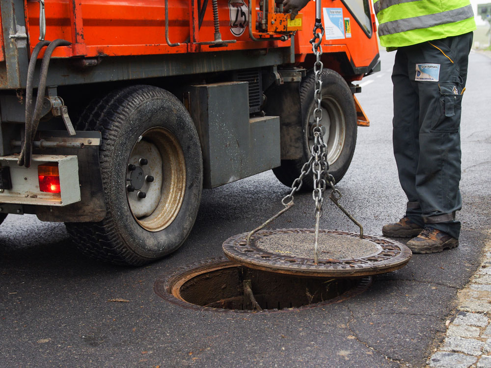 Ein Arbeiter in reflektierender Kleidung steht neben einem orangefarbenen Versorgungsfahrzeug und hebt mit einem Kettenmechanismus einen Gullydeckel von der Straße, so dass der darunter liegende offene Schacht sichtbar wird. Ein Arbeiter in reflektierender Kleidung steht neben einem orangefarbenen Versorgungsfahrzeug und hebt mit einem Kettenmechanismus einen Gullydeckel von der Straße, so dass der darunter liegende offene Schacht sichtbar wird.