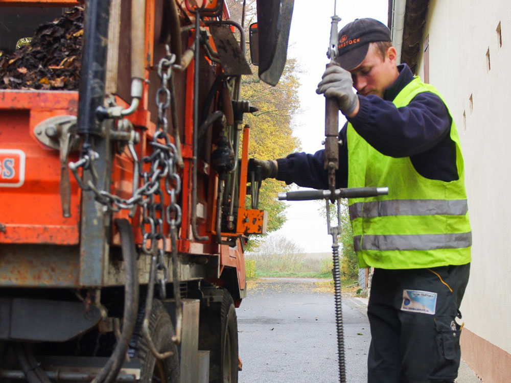 Ein Arbeiter in einer reflektierenden Weste bedient Maschinen neben einem orangefarbenen Versorgungsfahrzeug auf einer asphaltierten Straße. Er benutzt ein Metallwerkzeug, möglicherweise für Wartungs- oder Reparaturarbeiten, mit Herbstbäumen im Hintergrund. Ein Arbeiter in einer reflektierenden Weste bedient Maschinen neben einem orangefarbenen Versorgungsfahrzeug auf einer asphaltierten Straße. Er benutzt ein Metallwerkzeug, möglicherweise für Wartungs- oder Reparaturarbeiten, mit Herbstbäumen im Hintergrund.
