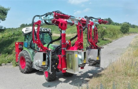 Ein grüner Traktor mit leuchtend roter Ausrüstung für die Weinbergsbeschneidung fährt auf einer asphaltierten Landstraße, umgeben von grasbewachsenen Hängen, Grünflächen und Weinbergsreihen unter blauem Himmel.