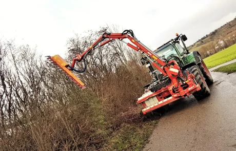 Ein grüner Traktor mit einer roten Heckenschere schneidet an einem bewölkten Tag zugewachsene Büsche am Rande einer Landstraße.