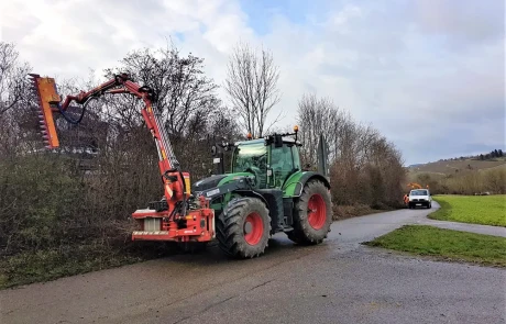 Ein Traktor mit einem Heckenschneidegerät schneidet Büsche entlang einer Landstraße. Ein anderes Fahrzeug ist weiter unten an der Straße in der Nähe von grasbewachsenen Feldern und kahlen Bäumen unter einem teilweise bewölkten Himmel geparkt.