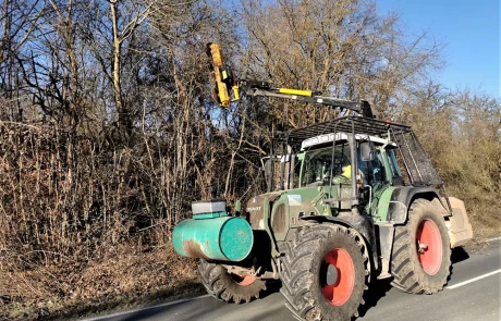 Ein grüner Traktor mit großen roten Rädern ist am Straßenrand geparkt, mit einem Metallarm und einem Maschinenaufsatz, der über dem Fahrerhaus angebracht ist. Laublose Bäume und Sträucher säumen die Straße unter einem klaren blauen Himmel.