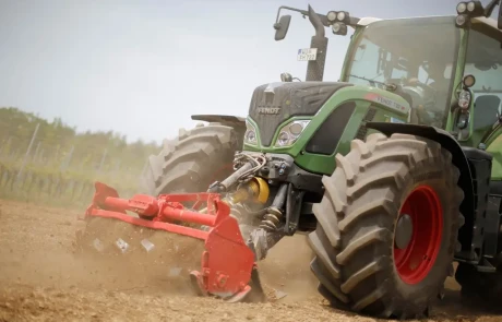 Ein grüner Fendt-Traktor mit großen Reifen pflügt mit einer roten Bodenfräse ein trockenes, staubiges Feld. Im Hintergrund sind Bäume und ein blauer Himmel zu sehen.